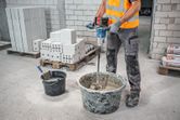 A person wearing safety equipment mixes mortar in a bucket with a stirring mechanism.