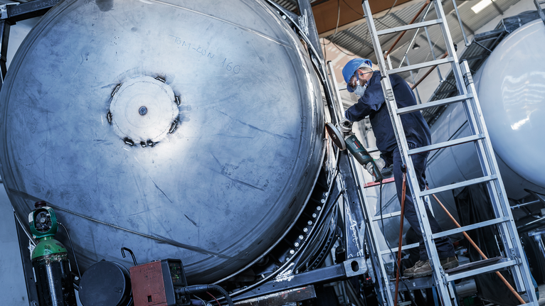 Person wearing safety equipment grinds the edge of a large metal tank on a ladder.