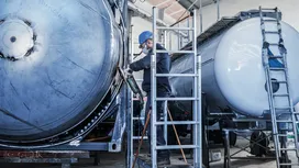 A person wearing safety equipment grinds a large metal tank with a power tool.