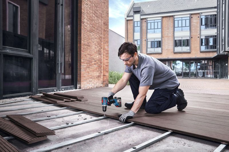 GSR 18V-65 A person wearing safety equipment uses a cordless drill to install decking boards outside.