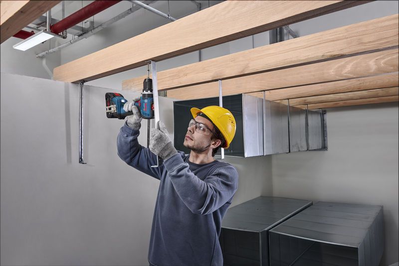 GSB 18V-65 A person wearing safety equipment drills into a metal rod beneath wooden beams.