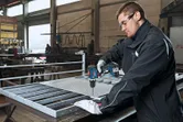A person wearing safety equipment drills metal mesh on a workbench in a workshop.