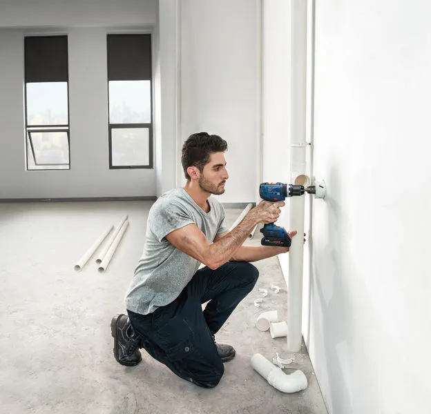GSB 18V-65 A person drills into a wall to install piping in a bright, unfinished room.