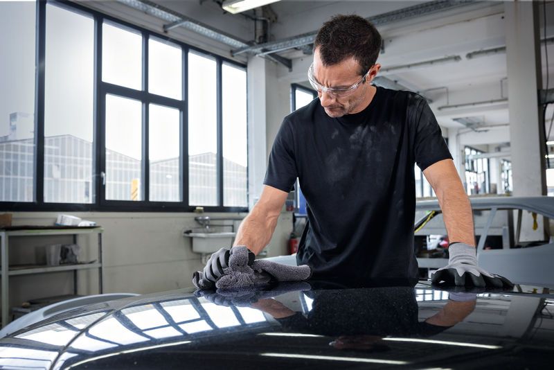 A person wearing safety equipment polishes a car hood with a cloth in a workshop.