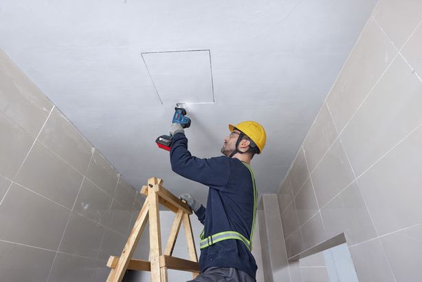 A person wearing safety equipment uses a reciprocating saw to cut a ceiling panel.
