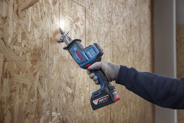 Person wearing safety equipment cuts wood panel using a cordless reciprocating saw.