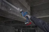 A person wearing safety equipment uses a reciprocating saw to cut metal overhead.