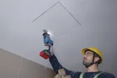 A person wearing safety equipment cuts a ceiling panel with a reciprocating saw.