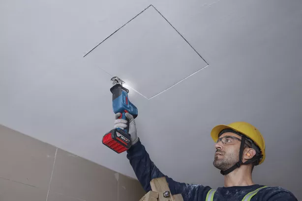 A person wearing safety equipment cuts a ceiling panel with a reciprocating saw.