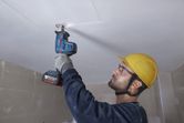 A person wearing safety equipment uses a reciprocating saw to cut into a ceiling panel.