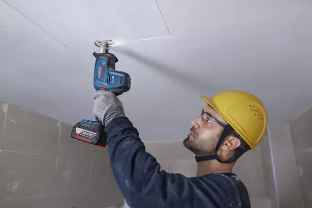 A person wearing safety equipment uses a reciprocating saw to cut into a ceiling panel.