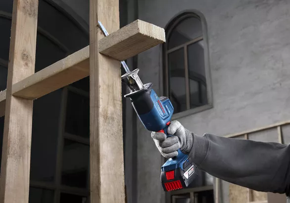 A person wearing safety equipment uses a reciprocating saw to cut wooden beams.
