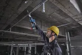 A person wearing safety equipment uses a reciprocating saw to cut a metal pipe overhead.