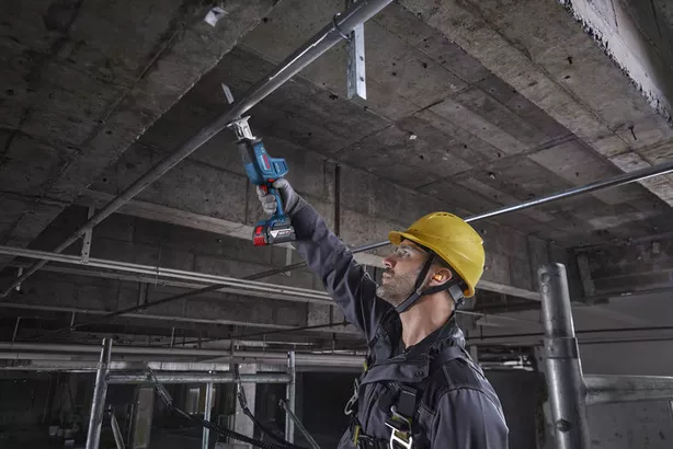 A person wearing safety equipment uses a reciprocating saw to cut a metal pipe overhead.