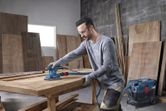 Person wearing safety equipment sands a wooden tabletop with an orbital sander.