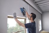 A person wearing safety equipment smooths a wall using an orbital sander near a window.