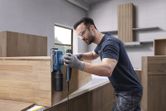 A person wearing safety equipment sands a wooden cabinet using an orbital sander.