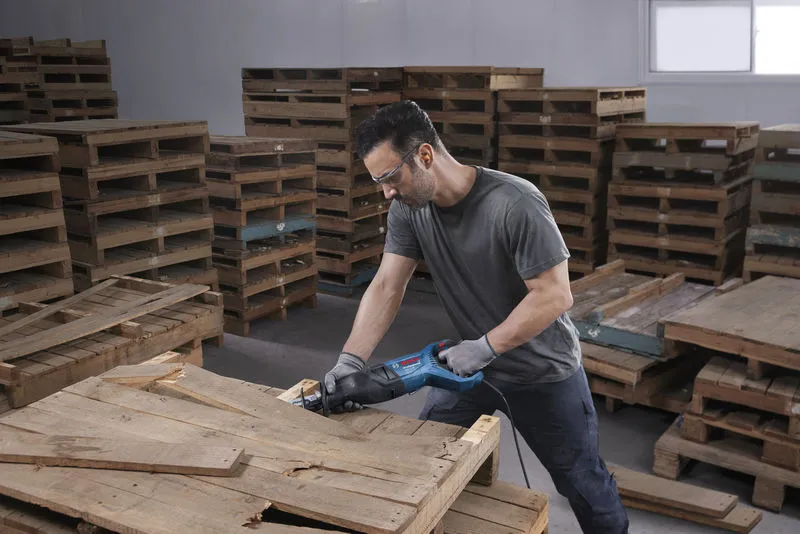 A person wearing safety equipment cuts a wooden pallet with a reciprocating saw.