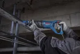 A person wearing safety equipment cuts a metal pipe with a reciprocating saw.