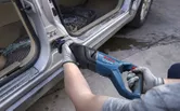 A person wearing safety equipment uses a reciprocating saw to cut metal on a car door frame.