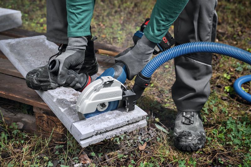 A person wearing safety equipment uses a large angle grinder with dust extraction to cut concrete.