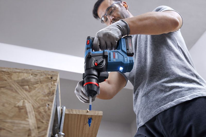 A person wearing safety equipment drills into wood with a cordless rotary hammer.