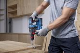 A person wearing safety equipment drills into wood using a cordless rotary hammer.