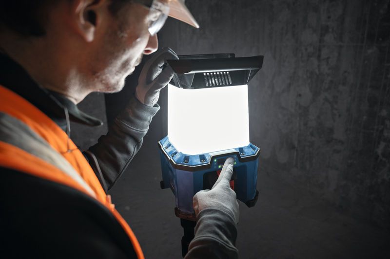 A person wearing safety equipment adjusts a cordless jobsite light in a dim workspace.