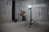 A person wearing safety equipment drills a concrete wall with a vacuum and standing jobsite light.