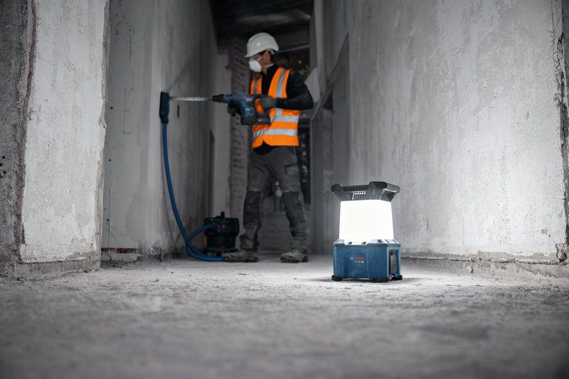 A worker wearing safety equipment drills a wall, cordless jobsite light on the floor.