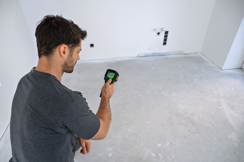 A person scans a bare room floor with a thermal camera.