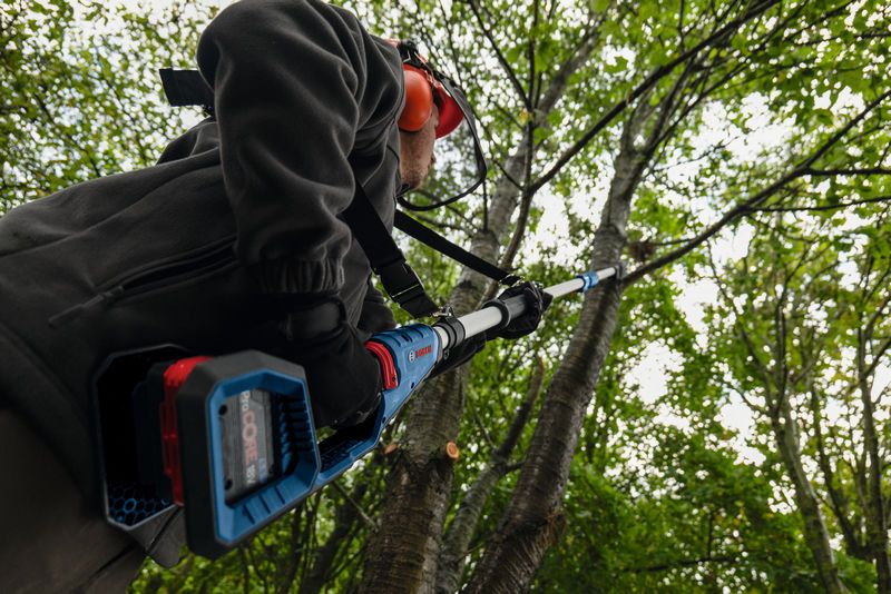 A person wearing safety equipment trims tree branches with an extendable power tool.