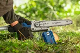 A person wearing safety equipment uses a chainsaw to cut a small tree branch outdoors.