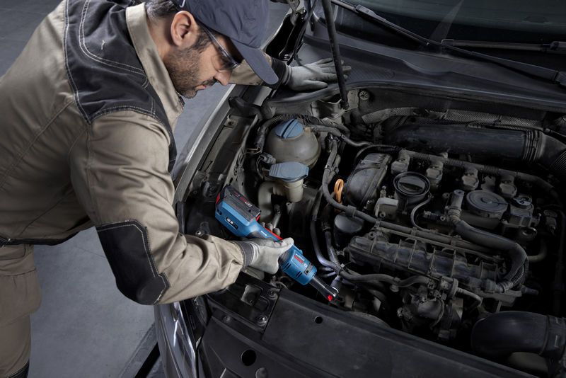 A person wearing safety equipment uses a cordless ratchet wrench to work on a car engine.