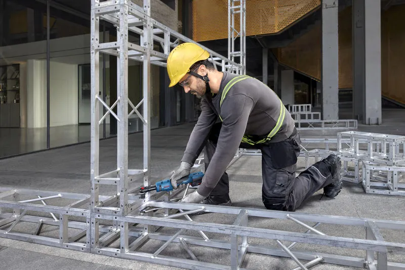 A person wearing safety equipment tightens bolts on a metal frame with a cordless ratchet wrench.