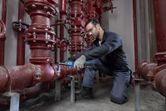 A person wearing safety equipment uses a cordless ratchet wrench on a red pipe system.