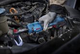 A person wearing safety equipment uses a cordless ratchet wrench to tighten a bolt in a car engine.