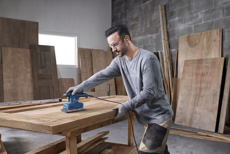 A person wearing safety equipment sands a wooden tabletop in a workshop.