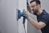 A person wearing safety equipment sands a wall with an orbital sander.