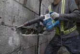 A person wearing safety equipment uses a demolition hammer to break a concrete wall.