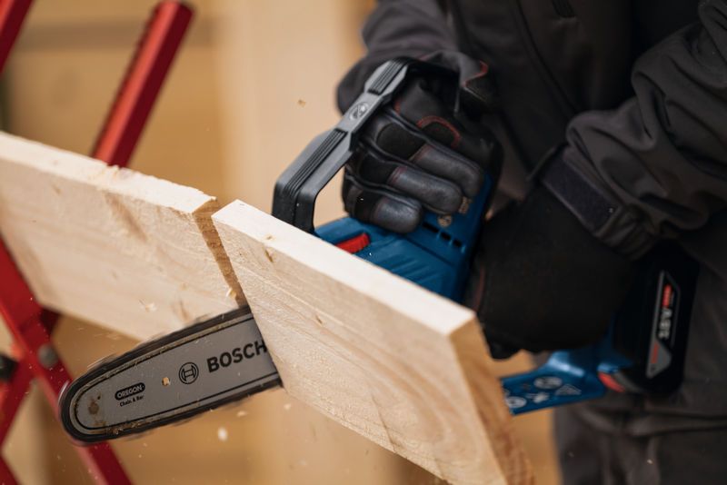 A person wearing safety equipment uses a cordless pruner saw to cut a wooden board.