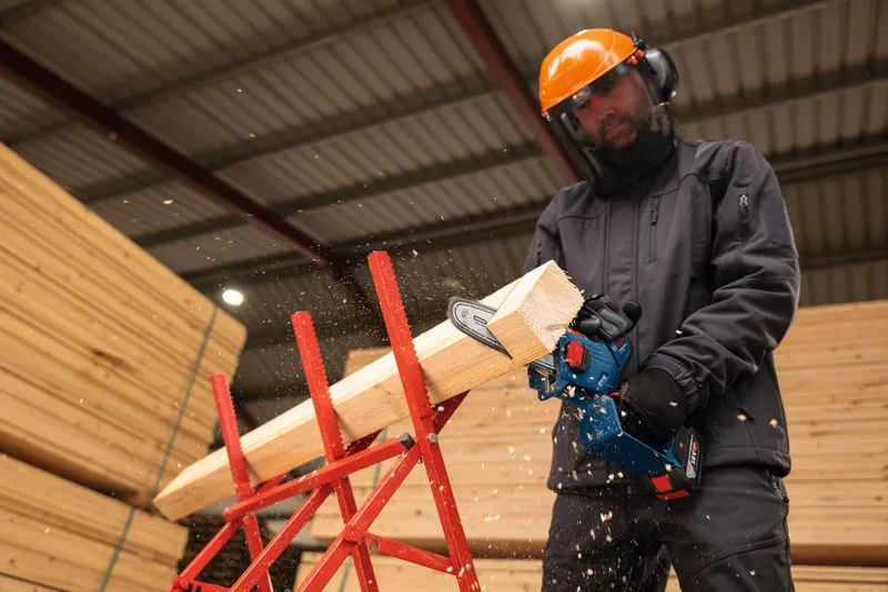 A person wearing safety equipment cuts lumber with a cordless pruner saw indoors.