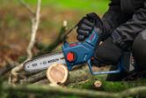 A person wearing safety equipment uses a cordless pruner saw to cut a tree branch.