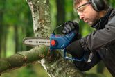 A person wearing safety equipment trims a tree branch with a cordless pruner saw.