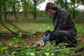A person wearing safety equipment trims branches on the ground with a cordless pruner saw.