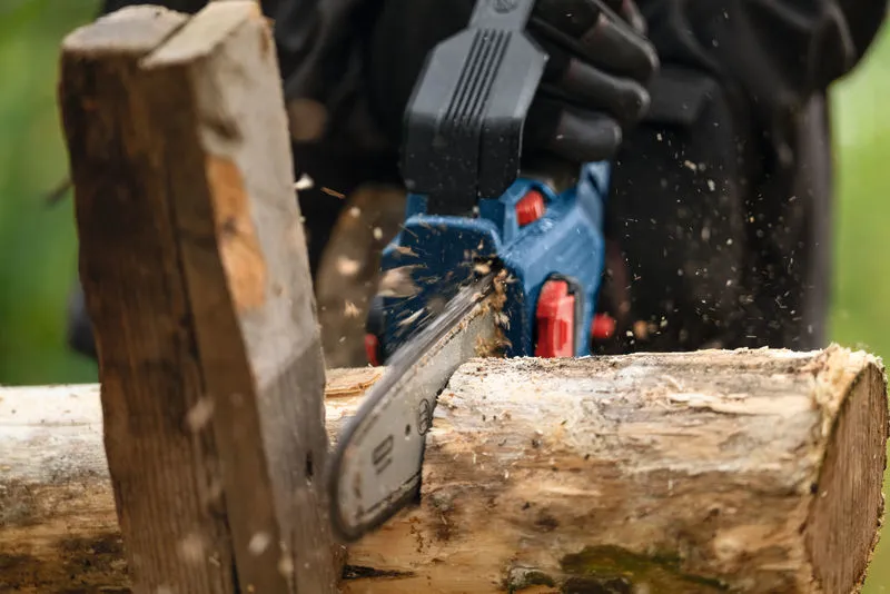 A person wearing safety equipment uses a cordless pruner saw to cut a log.