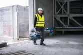 A person wearing safety equipment carries a metal cut-off saw in an unfinished building.