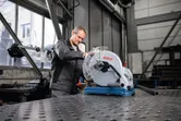 A person wearing safety equipment operates a metal cut-off saw in a workshop.