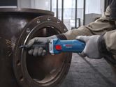 A person wearing safety equipment grinds a metal pipe with a straight grinder.