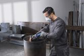 A person wearing safety equipment grinds the edge of a metal barrel with a straight grinder.